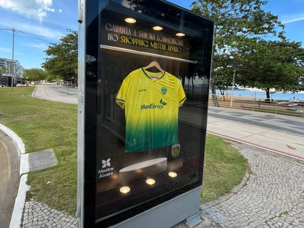 Vitrine em ambiente externo exibindo uma camisa amarela e verde do time Palmeiras, com logotipos e patrocinador, sobre iluminação interna e placas na fachada ao fundo com árvores e céu azul.