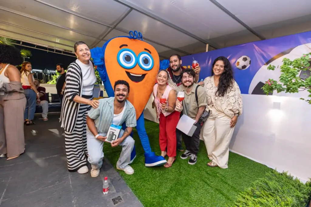 Grupo de pessoas em evento ao ar livre em área com grama sintética e mascote azul e laranja, celebrando com bebidas e brindes em ambiente decorado com tema esportivo.